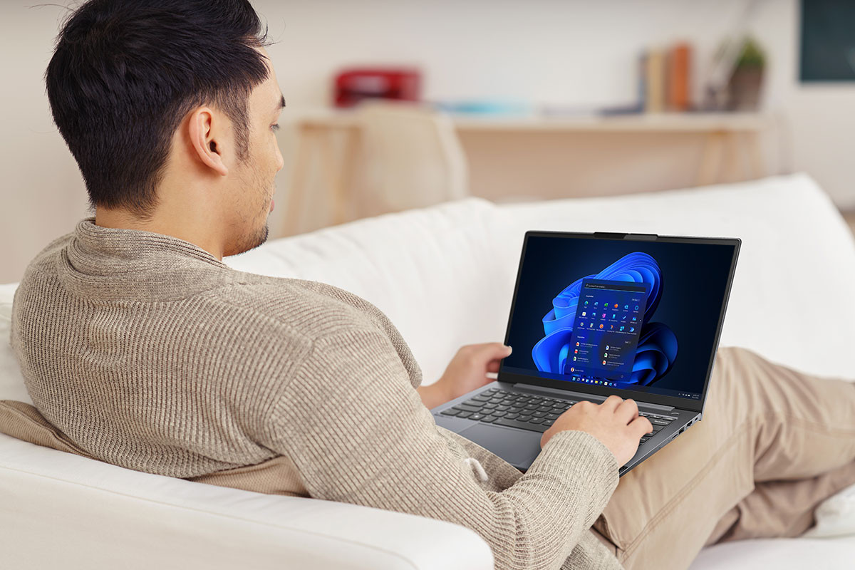 Ein junger Mann sitzt auf einer Couch und betrachtet sein Lenovo Yoga Laptop. Die Szene ist in warmen Beigetönen gehalten, was eine ruhige und gemütliche Atmosphäre vermittelt – Symbol für flexibles Arbeiten, Kreativität und modernes Home-Office.