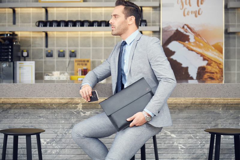 Eleganter Geschäftsmann in einem hellgrauen Anzug sitzt an einer modernen Café-Bar. Er hält eine Notebook Tasche in der Hand, während er nachdenklich zur Seite blickt.